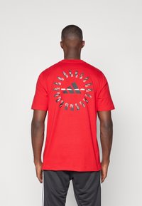 Camiseta de algodón roja con un gran logotipo negro de Adidas en la parte central de la espalda, rodeado de detalles gráficos blancos que asemejan zapatos.