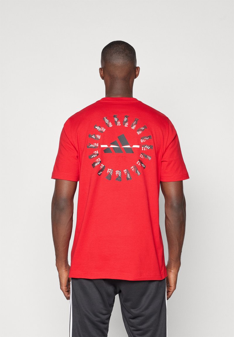 Camiseta de algodón roja con un gran logotipo negro de Adidas en la parte central de la espalda, rodeado de detalles gráficos blancos que asemejan zapatos.