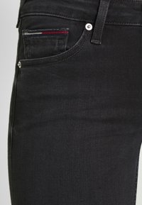 Pantalones vaqueros de mezclilla negra con corte ajustado, bolsillo frontal con detalles de costura en rojo y azul, y elementos de metal plateado en la cintura.