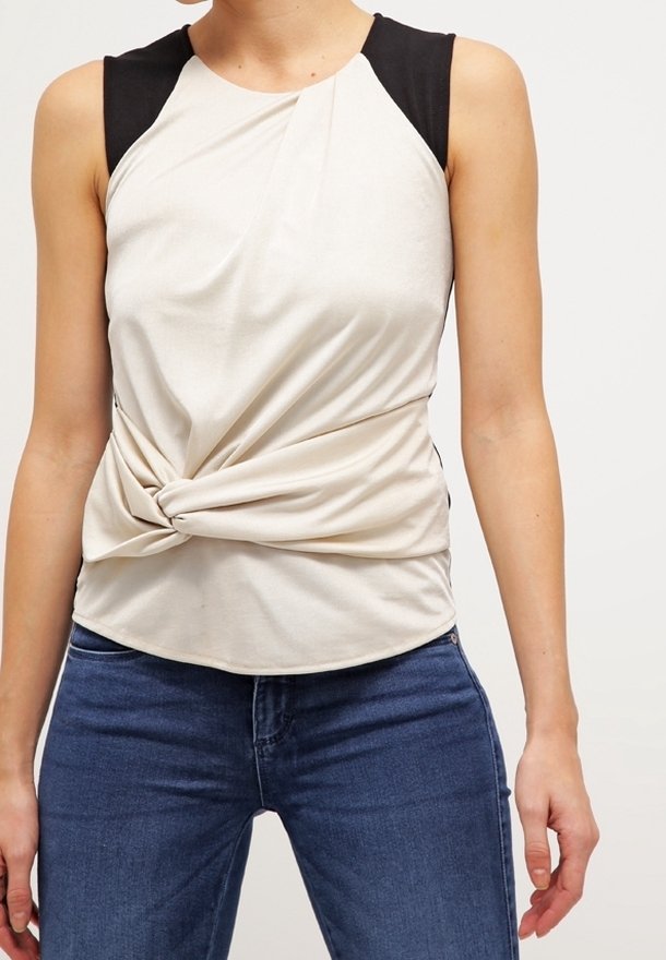 Ljusbeige ärmlös topp med svarta axeldetaljer, med en vriden frontdesign och mjukt tyg, parat med blå denimjeans.
