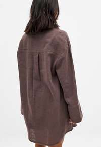 Camicia in lino a maniche lunghe di un marrone medio con una vestibilità rilassata, caratterizzata da una piega sul retro e un orlo arrotondato per una silhouette contemporanea.
