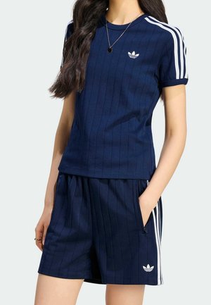 Kobieta ubrana w granatową koszulkę Adidas z krótkim rękawem i pasujące szorty z białymi paskami i logo, ręka w kieszeni.