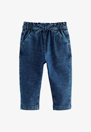 Młoda dziewczyna z blond włosami związanymi w kucyki, ubrana w szary golf, niebieskie dżinsy i białe buty na rzepy, stojąca wewnątrz pomieszczenia.