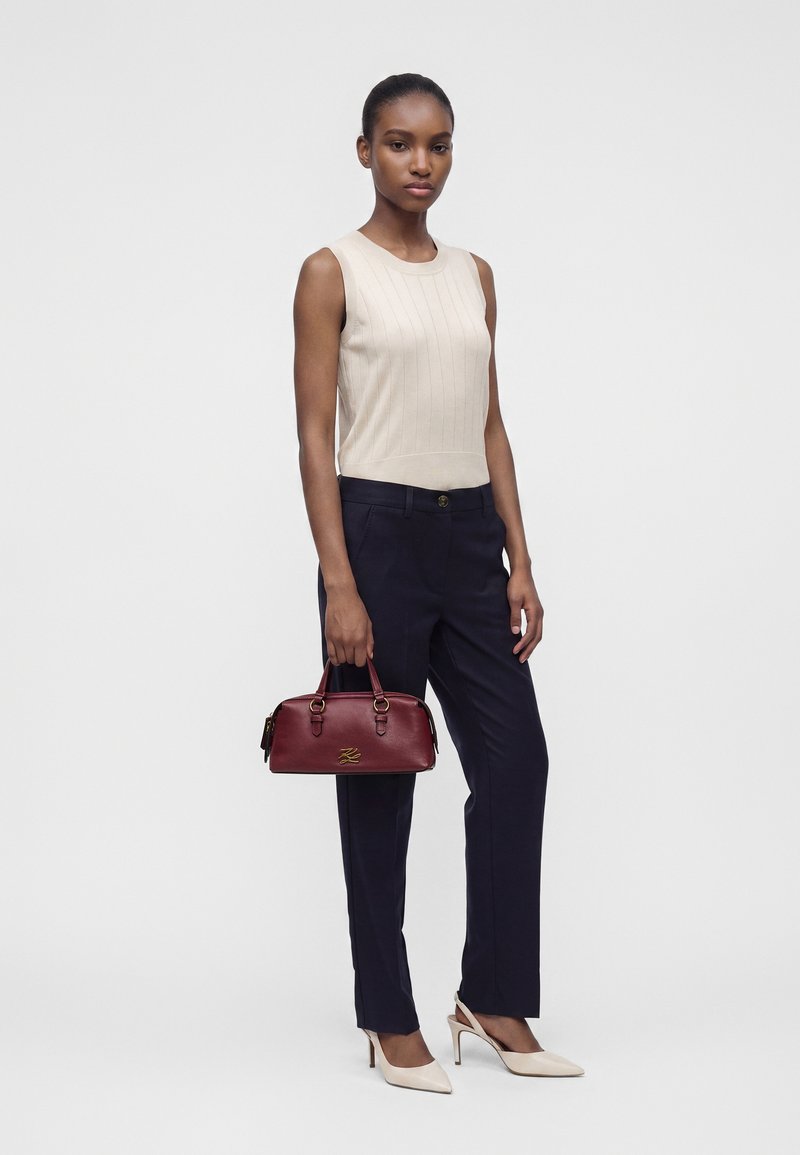 Γυναίκα που φοράει κρεμ αμάνικη μπλούζα, μαύρο παντελόνι, κρεμ τακούνια, κρατώντας μικρή μπορντό τσάντα, στέκεται μπροστά σε απλό φωτεινό φόντο.