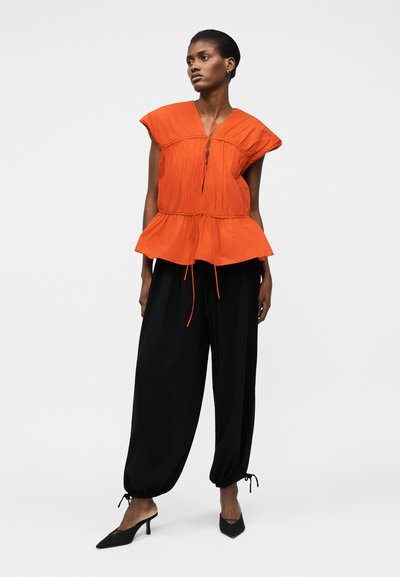 Naine, kes seisab mustades laiade säärega pükstes, millel on paelad, ning oranžis varrukateta topis, millel on lehviva serva ja ees paelad, kandes musti kontsakingi.