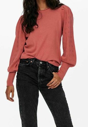 Vrouw in een stofroze lange mouwen trui met pofmouwen en zwarte hoge taille jeans, met één hand in de zak.