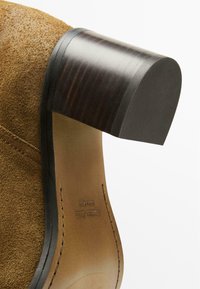 Stivaletto alla caviglia in suede marrone con tacco a blocco in legno nero lucido. Presenta un bordo del tacco arrotondato e cuciture visibili sulla suola.