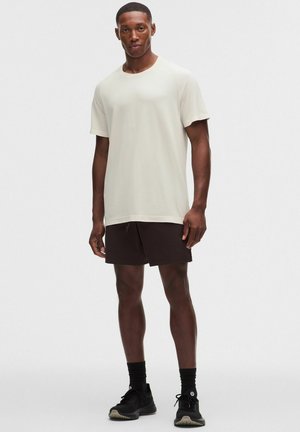 Mand iført en lys beige kortærmet T-shirt, sorte shorts, sorte sokker og sorte sportssko, stående mod en ensfarvet baggrund.