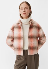 Różowo-pomarańczowa kurtka w kratkę o miękkiej teksturze, z kołnierzykiem i zatrzaskami, noszona na kremowym swetrze i beżowych spodniach.