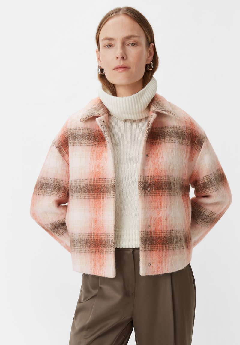 Różowo-pomarańczowa kurtka w kratkę o miękkiej teksturze, z kołnierzykiem i zatrzaskami, noszona na kremowym swetrze i beżowych spodniach.