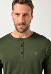 Mies, jolla on lyhyt hius ja vaalea parta, pukeutuneena oliivinvihreään henley-paitaan, jossa on kolme mustaa nappia ja huomaamaton "JP 1880" -logo rinnassa.