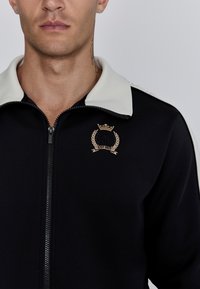Veste noire zippée avec un col blanc, présentant un logo et des détails brodés en or. Tissu lisse avec une coupe moderne.