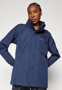 Navy waterproof jacket with a high collar, two front pockets, and elastic cuffs. Features branding on the chest and left sleeve. Smooth texture.