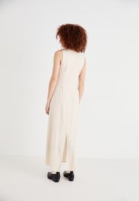 Robe en lin beige sans manches avec une coupe droite, des plis au dos et une fente haute. Portée avec des chaussures noires avec des accents crème.