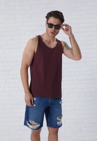 Burgundska majica bez rukava od pamuka, u kombinaciji s iznošenim plavim trapericama s više otvora. Model nosi crne sunčane naočale.