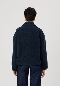 Granatowa, miękka, strukturalna kurtka z polaru z wysokim kołnierzem i luźnym krojem, posiadająca długie rękawy i przeszycia z tyłu.