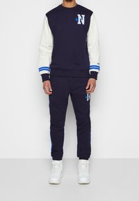 Navy sweatshirt with white sleeves, blue accents, and an embroidered logo. Coordinating navy joggers feature blue side stripes. Worn with white sneakers.