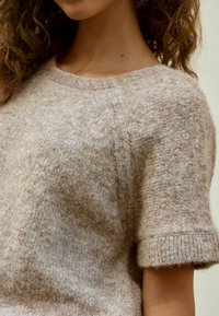 Beżowy sweter dziany z krótkim rękawem, miękka tekstura, okrągły dekolt i subtelne tonalne zróżnicowania. Posiada ściągacze przy mankietach i u dołu.