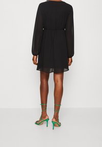 Robe noire à manches longues avec une taille cintrée, réalisée dans un tissu léger. Associée à des sandales à talons hauts vertes à lanières.