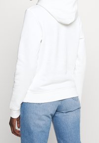 Felpa bianca con cappuccio, tasca a marsupio, polsini e orlo a costine. Abbinata a jeans denim azzurri chiari, che mostrano un fit rilassato.