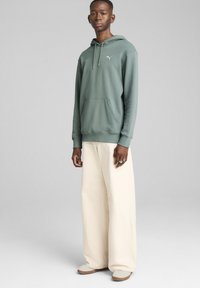 Grøn bomulds hoodie med en frontlomme og snoretræk ved hætten, kombineret med løstsiddende cremefarvede bukser og lyse sko.