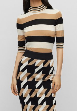 Nainen, jolla on beige, musta ja valkoraidallinen lyhythihainen paita ja siihen sopiva geometrinen kuvioitu hame yksiväristä taustaa vasten.