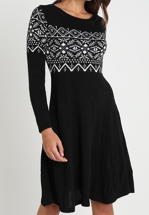 Robe noire à manches longues en maille avec des motifs géométriques et floraux blancs sur la poitrine et le haut des manches, corsage ajusté et jupe évasée.