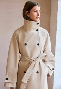 Beige dobbeltradet trench coat med store sorte knapper, bred krave og matchende bælte, der har en glat tekstur og afslappet pasform.
