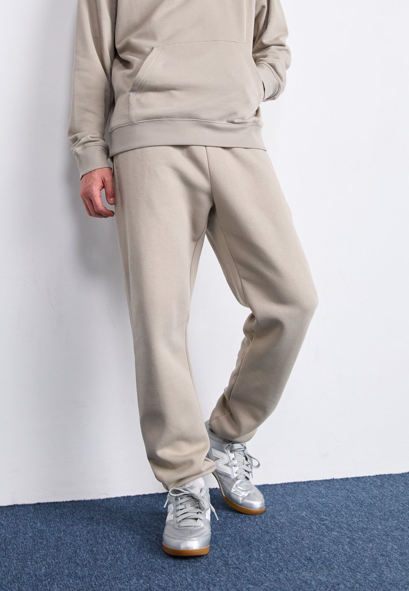 Beige Sweatshirt mit Kängurutasche und passenden Jogginghosen aus weichem Stoff; kombiniert mit silbernen Sneakers mit hellgrauen Schnürsenkeln.