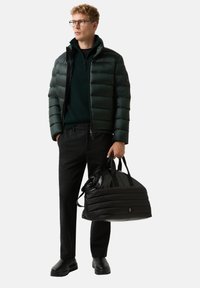 Junger Mann mit Brille, schwarzer Steppjacke, dunkler Hose und Stiefeln, der eine große schwarze gepolsterte Reisetasche an den Griffen hält.