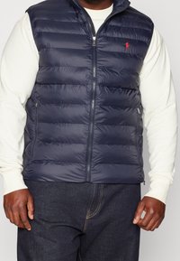 Man med marinblå vadderad zip-up väst över en vit långärmad skjorta och mörkblå jeans, med händerna avslappnat vid sidorna.