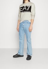 Ljust grå sweatshirt med svart och vitt grafiskt logo, tillsammans med ljusblå denimjeans och svarta sneakers. Avslappnad, lös passform.