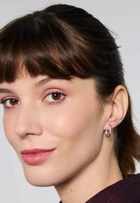 Srebrny kolczyk w formie obręczy o gładkim, eleganckim wzornictwie. Modelka ma średniej długości ciemne brązowe włosy i delikatny makijaż.