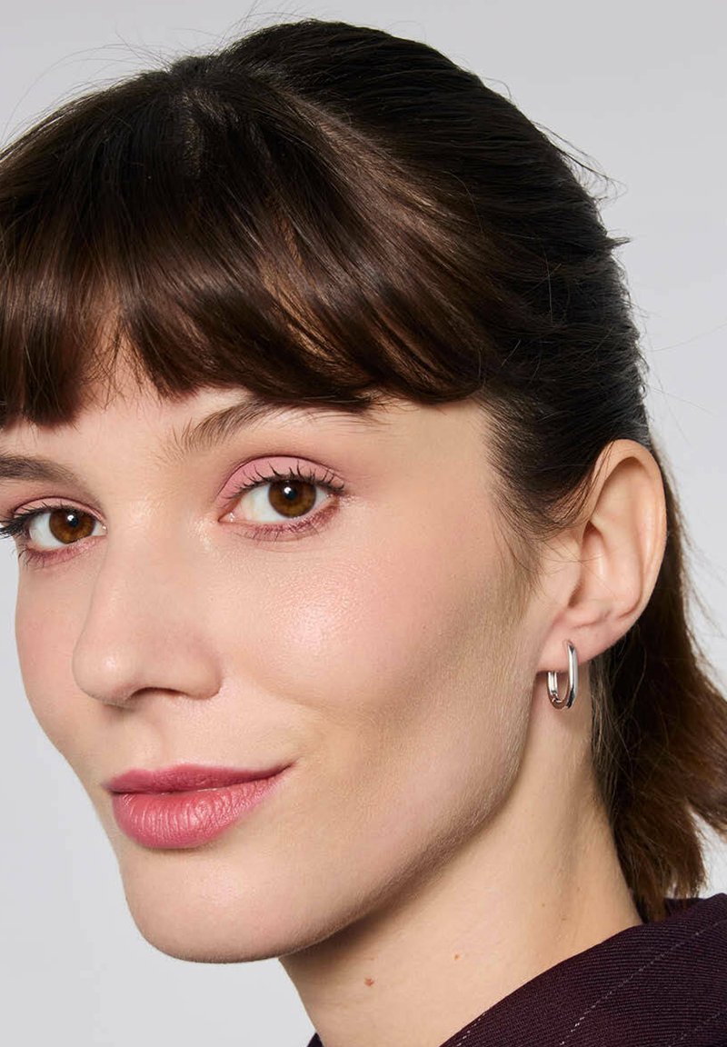 Srebrny kolczyk w formie obręczy o gładkim, eleganckim wzornictwie. Modelka ma średniej długości ciemne brązowe włosy i delikatny makijaż.