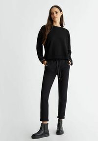 Felpa nera con maniche testurizzate, abbinata a pantaloni neri con coulisse e tasche laterali, dotati di dettagli in metallo color oro. Stivali neri alla caviglia.