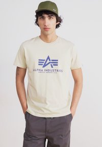 Beige bomulds t-shirt med et blåt trykt logo, rund halsudskæring og korte ærmer, parret med grå bukser og en grøn kasket.