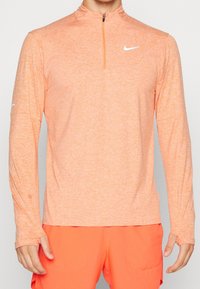 Mies, který má na sobě dlouhý rukáv, světle oranžové sportovní tričko Nike a jasně oranžové šortky s otvory na palce ve rukávech.