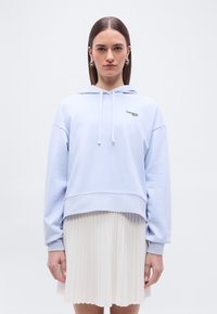 Femme portant un sweat à capuche bleu clair avec cordons et un petit logo, associée à une jupe plissée blanche, debout devant un fond uni.