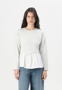 Top gris de manga larga con un peplum blanco fruncido. La textura del tejido suave y las capas contrastantes crean una silueta relajada. Se lleva con jeans azules.