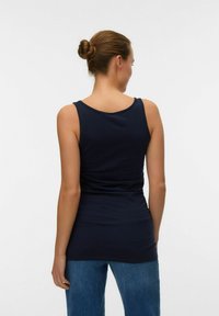 Kobieta stoi tyłem do kamery, ubrana w granatowy bezrękawnik i niebieskie dżinsy, z włosami związanymi w kok na białym tle.