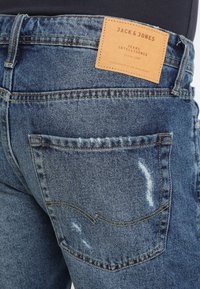 Džínové kalhoty středně modré barvy s opotřebovaným vzhledem, s koženou značkou na pasu a zadními kapsami se švy.