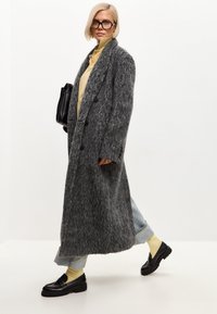 Abrigo largo de lana gris con un patrón texturizado, que presenta grandes botones y un corte relajado. Combinado con una camisa amarilla de cuello y jeans.