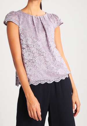 Vrouw met een lichtpaarse kanten blouse met korte mouwen en golvende zoom, en een zwarte broek, staand tegen een egale achtergrond.