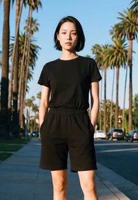 Ung kvinde med kort sort hår iført sort t-shirt og shorts, der står på en gade med palmetræer og parkerede biler.