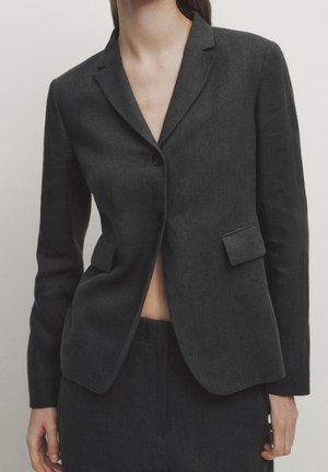 Femme portant un blazer cintré gris foncé à deux boutons et un pantalon assorti, sur un fond clair uni.