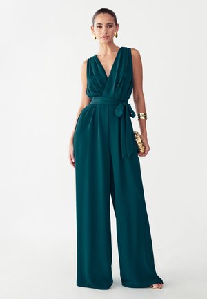Kvinde iført ærmeløs grønblå jumpsuit med vide ben, dyb V-hals og knyttet talje, accessoriseret med guldringe, armbånd og clutch.