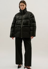 Černá puff jacket s vysokým límcem, která má zipové zapínání a prošívanou texturu. V kombinaci s volnými černými kalhotami a špičatými podpatky.