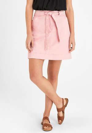 Femme portant une jupe rose ceinturée, un haut noir et des sandales plates marron, debout, jambes croisées, sur un fond blanc.