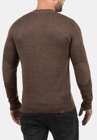 Brun strikkegenser med lange ermer, ribbestrikkede mansjetter, albueflater og rund hals; jevn tekstur og tettsittende silhuett.