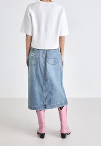 Haut blanc à manches courtes plissé, jupe midi en denim bleu avec poches arrière, et bottes de cow-boy roses, vues de derrière sur un fond neutre.